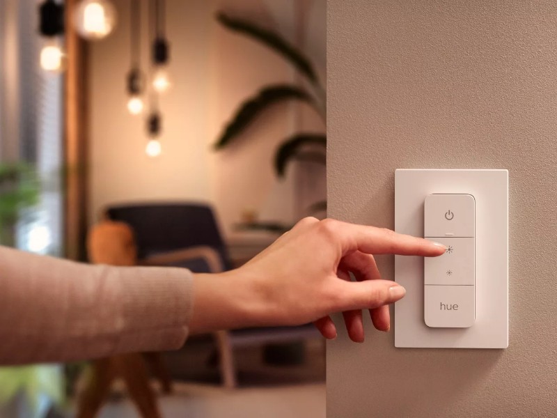 Hand drukt op witte Hue Dimmer Switch op beige muur om licht feller te zetten. Wazige woonkamer met warm gloeiende lampen op de achtergrond.