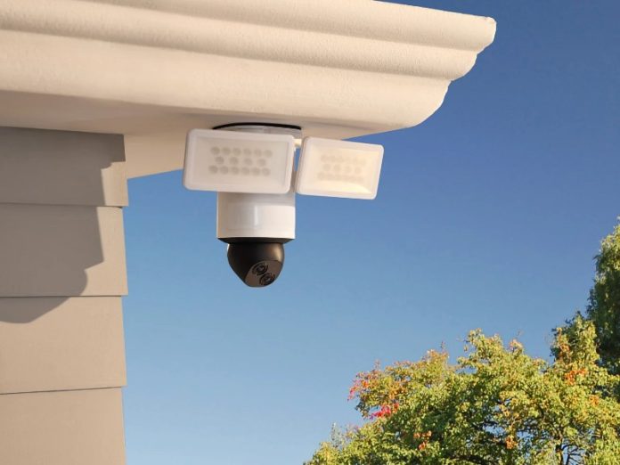 Witte beveiligingscamera met twee LED-schijnwerpers en een zwarte bolvormige camerakop met dubbele lens, gemonteerd onder een dakrand tegen een blauwe lucht met bomen.