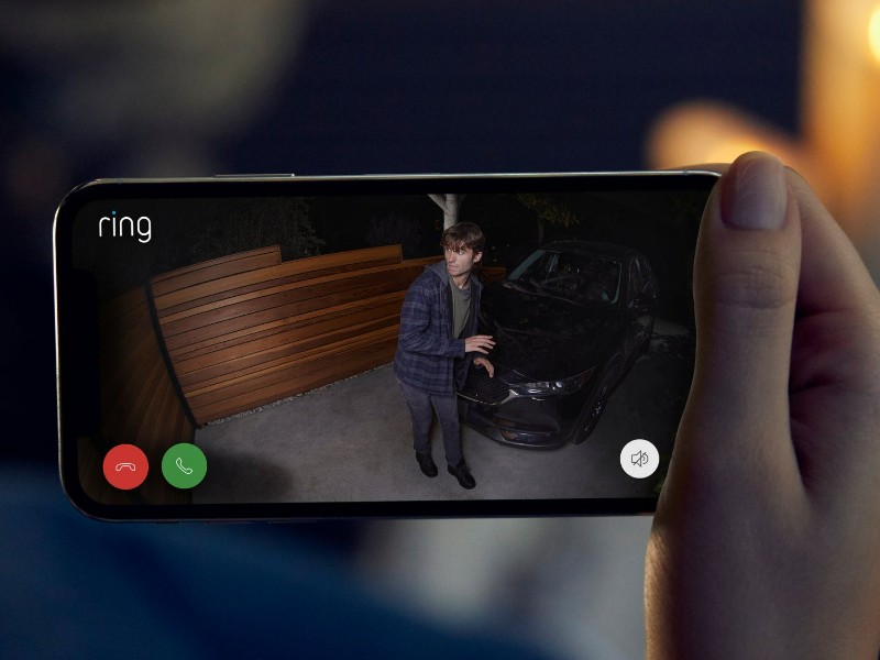 Een hand houdt een smartphone vast met daarop live camerabeelden van een Ring-systeem. Op het scherm is 's nachts een verdachte man bij een donkere auto te zien.