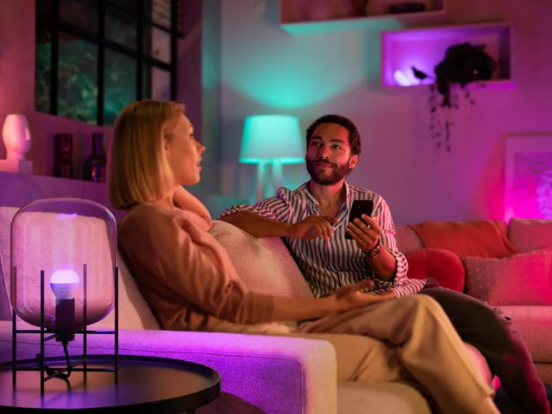 Image eines Paares in einem smart beleuchteten Wohnzimmer. Pinke, lila und türkise Lichter erzeugen eine stimmungsvolle Atmosphäre. Der Mann hält ein Smartphone.