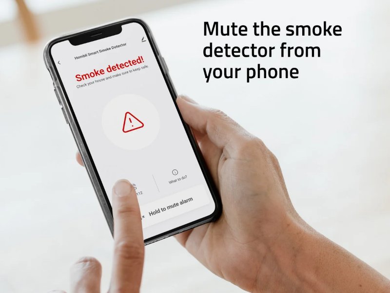 Een hand houdt een smartphone vast die een "rook gedetecteerd"-melding toont met een waarschuwingsdriehoek en de optie om het alarm te dempen.