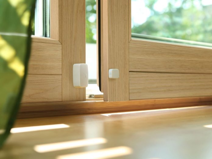 Een witte slimme raam- of deurcontactsensor, bestaande uit twee gescheiden delen, gemonteerd op een lichtbruin houten kozijn.