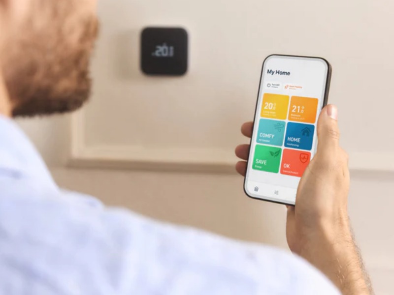 Een persoon houdt een smartphone vast waarop een slimme-app wordt getoond, met op de achtergrond een slimme thermostaat.