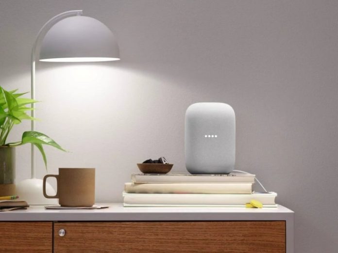 Een Google Nest smart speaker staat op een stapel boeken op een nachtkastje, met een plant en een bureaulamp ernaast. Een tekstballon op de muur toont de opdracht 'Hey Google, turn on the lights', wat de spraakbesturingsfunctie van de slimme luidspreker illustreert.