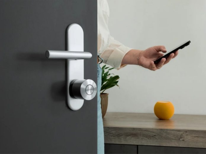Een slim deurslot op een donkere deur en een hand die een smartphone vasthoudt voor bediening, naast een slimme luidspreker.
