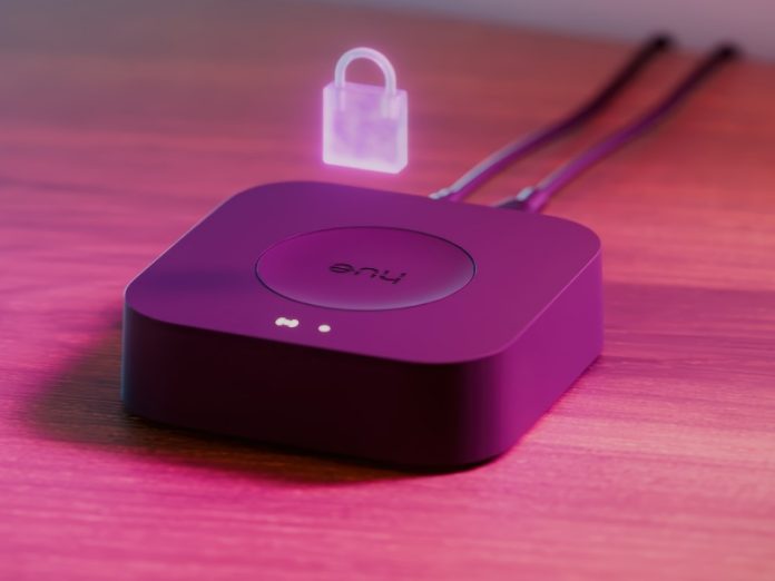 Philips Hue Bridge Pro op een houten oppervlak, verlicht met roze-paars licht, met een gloeiend hangsloticoon erboven en twee witte statuslampjes.