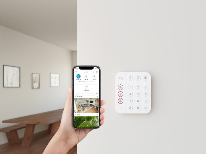 ring_vs_eufy-7 Een hand toont de Ring app op een smartphone met camerabeelden van een woonkamer en tuin, naast een Ring alarmsysteem keypad aan de muur, relevant voor Ring vs eufy vergelijkingen.