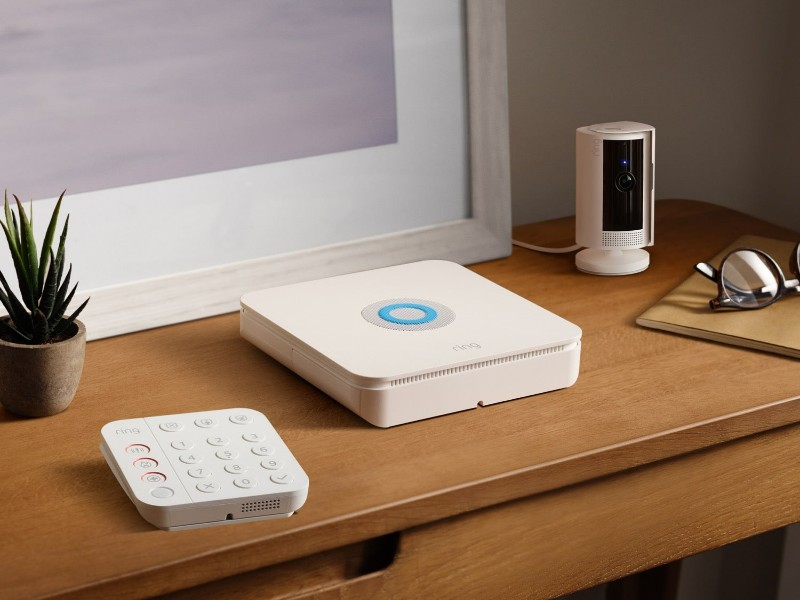 Een witte Ring beveiligingscamera, basisstation en keypad op een houten bureau. Dit Ring-systeem toont opties voor huisbeveiliging in de context van Ring vs eufy.