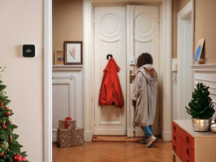 Moderne thermostaat toont 20.0 graden in een warm, kerstversierd huis. De Stadsverwarming zorgt voor een comfortabele temperatuur terwijl iemand de voordeur opent.
