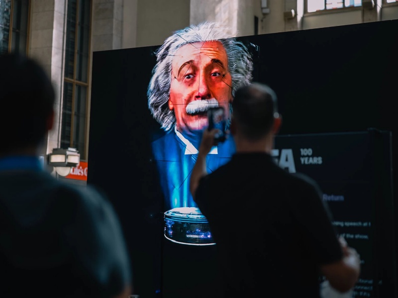 Digitale projectie van Albert Einstein op een groot scherm, bekeken en gefotografeerd door bezoekers. De installatie met een futuristisch rond apparaat toont de mogelijkheden van Ambient AI voor boeiende interactieve publieke ervaringen.