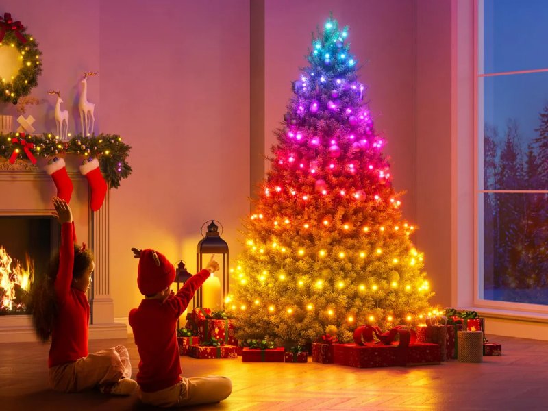 Twee kinderen zitten op de vloer in een sfeervolle kerstkamer, kijkend naar een grote kerstboom met 'Christmas Sparkle String Lights' die in regenboogkleuren van blauw bovenaan naar geel onderaan oplichten. Links bevindt zich een brandende open haard, versierd met een groene slinger, rode kerstsokken en een krans. Rechtsonder liggen cadeautjes en een raam biedt uitzicht op een besneeuwd winterlandschap.