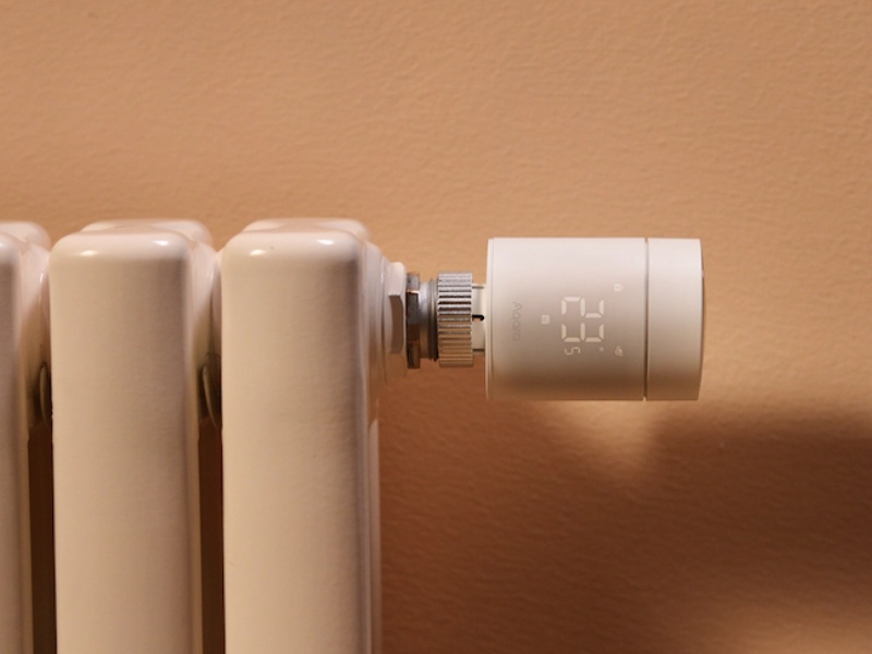 De W600 Thermostaat, een witte slimme thermostaat met digitaal display ('23.5') en 'Aqara' erop, bevestigd aan een witte radiator tegen een lichtbruine muur.