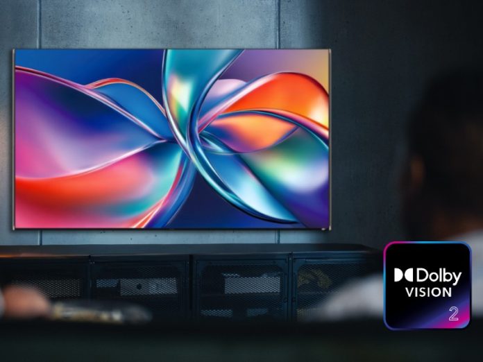 Twee onscherpe figuren bekijken een tv-scherm aan een donkergrijze muur, waarop een abstracte weergave van vloeiende, iriserende vormen in fel blauw, paars, oranje en rood te zien is, met rechtsonder het Dolby Vision 2-logo.