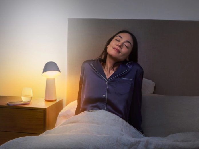 Vrouw in donkerblauwe satijnen pyjama zit ontspannen met gesloten ogen in bed, verlicht door de warme, oranje gloed van een witte en lichtblauwe paddestoellamp. Op het nachtkastje staat de lamp naast een glas water en een paarse smartphone, een serene setting voor het einde van de zomertijd en het begin van de wintertijd.