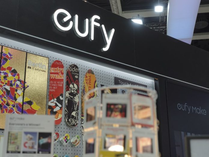 Gloeiend wit 'eufy' logo bovenaan een donkere beursstand. Links hangen kleurrijke mozaïek kunstwerken van een stadskaart en drie skateboards met grafische prints (o.a. rood en zwart), met daaronder diverse mokken. Een bord vermeldt 'Touch And Feel!' en een donker paneel toont 'eufy Make'. Een levendige sfeer op de Eufy IFA 2025 beursstand.