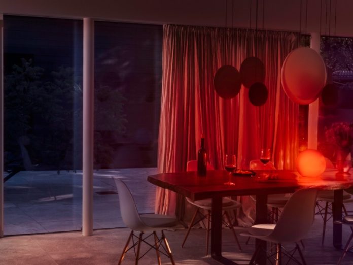 Eve Flare in Herfst: een moderne eetkamer in de avond met een warme, oranjerode sfeer. Een gloeiende bolvormige lamp verlicht een houten tafel met rode wijn en witte stoelen. Links reflecteert een donker terras met bomen in grote glazen schuifdeuren, rechts hangen roodbelichte gordijnen.