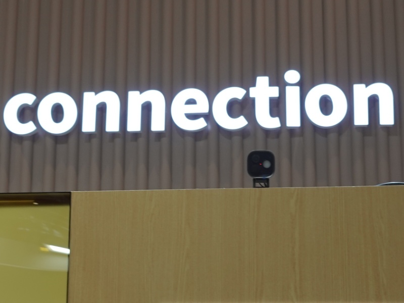 Een Aqara smart home productpresentatie op een beursstand. Op een beige geribbelde wand staat in witte, lichtgevende letters 'continuous connection'. Daaronder een display met diverse witte Aqara smart home producten, zoals camera's (inclusief de G510), sensoren en deursloten, op een geel verlichte achtergrond. Rechts hiervan toont een houten paneel een oranje bord 'Aqara Hubs' met informatie over Matter controllers, gevolgd door een sectie over 'Third-Party' compatibiliteit.