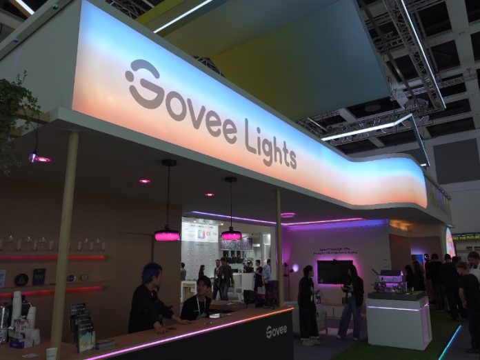 Govee IFA 2025: een levendige stand met een groot, lichtgevend 'Govee Lights' bord in een blauw-oranje kleurovergang. De stand is verlicht met diverse roze en paarse LED-accenten, met bezoekers die producten bekijken bij de balie en displays.