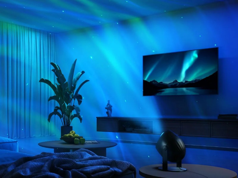 Sfeervolle woonkamer met blauwgroene aurora- en sterrenprojecties op muren en plafond, afkomstig van de Govee Star Light Projector op een bijzettafel. Een donkere bank, plant en tv vullen de ruimte.