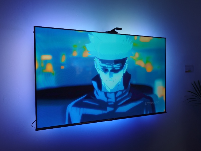 Een tv toont een blauw/cyaan anime-personage, terwijl de Govee TV Backlight 3 Pro de omringende muur in bijpassend paarsblauw licht dompelt. Rechts staat een plant in silhouet.
