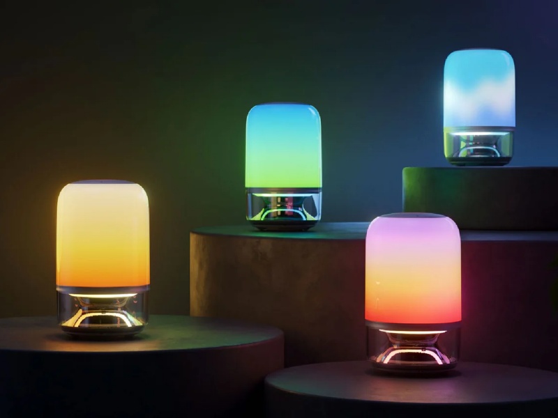 Vier Govee x JBL Table Lamp 2 Pro lampen, elk met een transparante onderkant, staan op donkere platforms. Ze gloeien in unieke kleurverlopen: geel-oranje, groen-lichtblauw, paars-oranje en lichtblauw-wit, tegen een schemerige achtergrond met een vaag zichtbare groene plant.