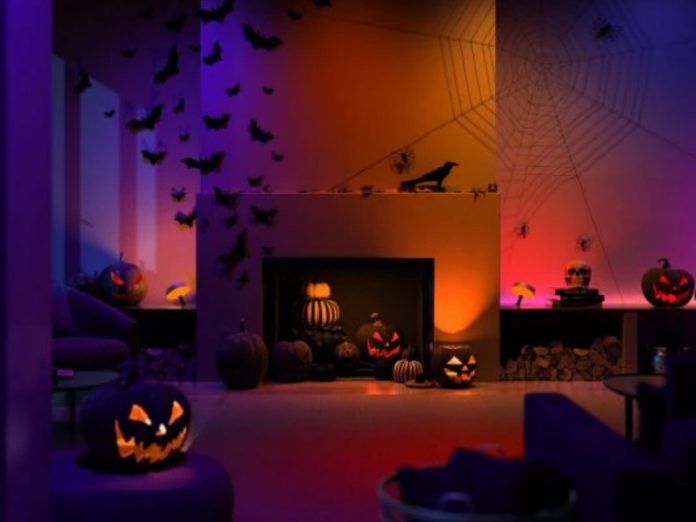 Een Halloween smart home kamer sfeervol verlicht met dynamische paarse, oranje, rode en blauwe tinten, toont zwarte vleermuizen op de linkermuur en grote spinnenwebben met spinnen op de rechter. Griezelig brandende pompoenen, waaronder een grote vooraan links, een zwarte kraai en schedels decoreren de ruimte.