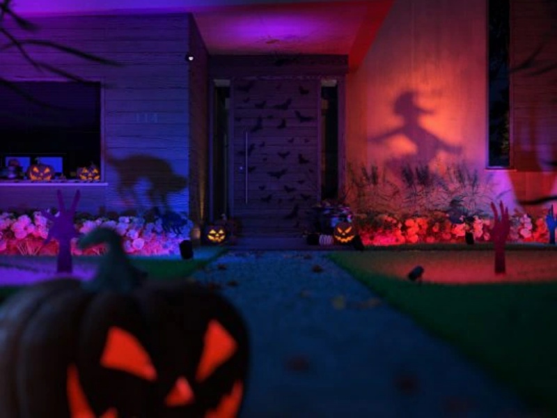 Een modern Halloween smart home 's nachts, levendig verlicht met paarse en rode lampen. Een heksenschaduw is geprojecteerd op een rode muur en een kat op een blauwe. Gloeiende pompoenlantaarns, waarvan één wazig op de voorgrond, en vleermuizen op de deur. Zombiehanden steken uit kleurrijke bloembedden.