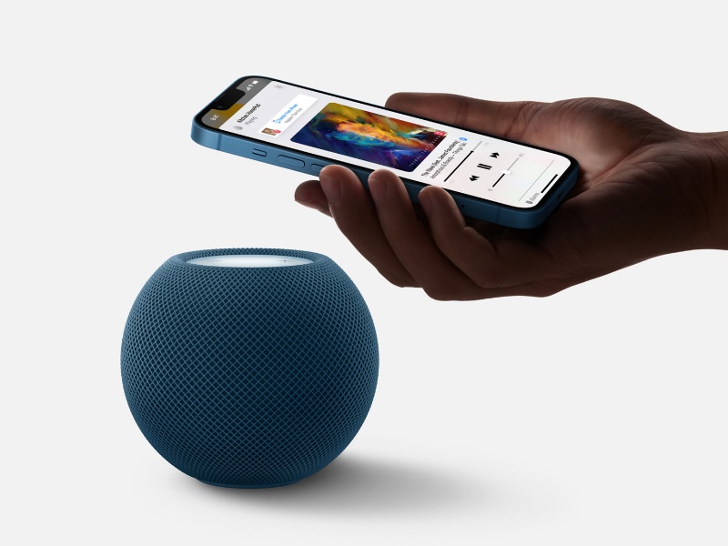 Een hand houdt een lichtblauwe iPhone boven een middernachtblauwe HomePod mini met gaastextuur en een oplichtende witte bovenkant. Het iPhone-scherm toont de tekst 'KitchenHomePod playing' met actieve muziekbediening en albumhoezen.