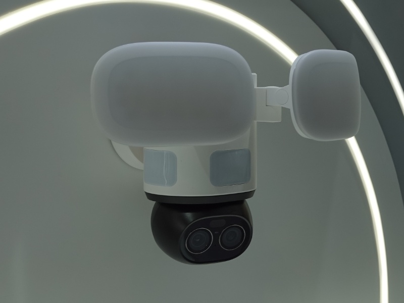 De Reolink Flex Floodlight WiFi beveiligingscamera, wit met een zwarte lensbehuizing en twee witte lichtpanelen, gepresenteerd op een stijlvolle, wit-grijze stand met gebogen LED-verlichting, zoals te zien op Reolink IFA 2025. Op de achtergrond vage beursbezoekers en een blauwe wand.