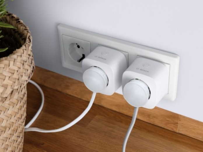 Twee witte Bosch slimme stekkers met energiemeting in een wit stopcontact, tegen een witte muur boven een houten plint. Links staat een rieten bloempot met groene plant.