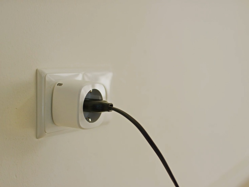 Een witte slimme stekker met energiemeting, aangesloten op een crèmekleurige muur, met een zwarte kabel erin.