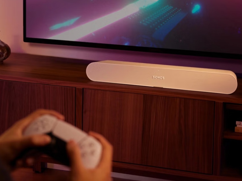 In een Smart Gaming Room speelt iemand een videogame met een witte controller. Een lichtbeige Sonos soundbar staat onder een tv die een paarse gamescène toont, op een donkerhouten meubel.