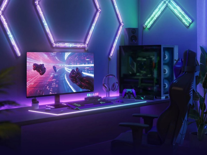 Een donkere 'Smart Gaming Room' met paarse, blauwe en groene neon-LED-verlichting. Op een bureau staat een gaming-pc met monitor die een futuristisch spel toont, een verlicht toetsenbord, koptelefoon en paarse controller. Paarse zeshoekige en groene V-vormige LED-lampen sieren de muren, met een gamingstoel en kamerplant ernaast.
