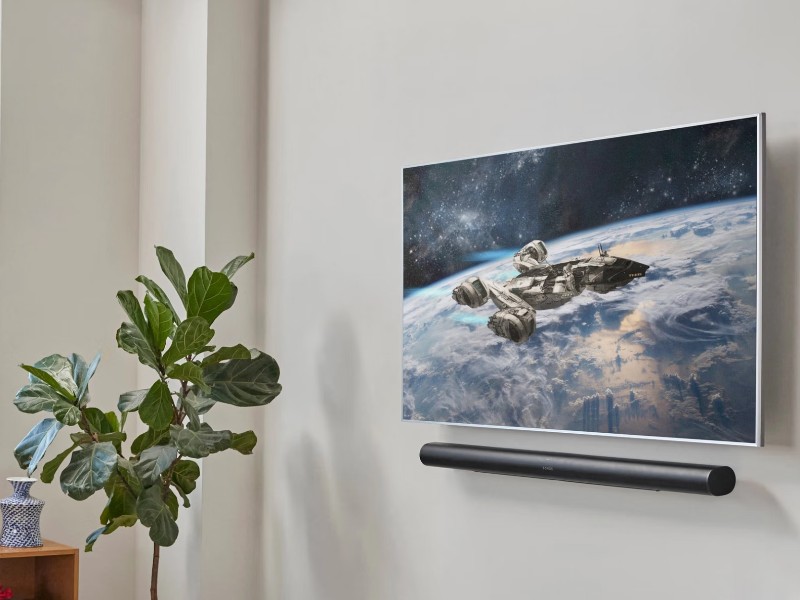 Moderne woonkamer met een groot raam links. Op de witte muur hangt een tv met een ruimteschip boven de aarde, daaronder een zwarte Sonos Arc Ultra soundbar. Links een grote groene vijgenbladplant in gele pot, naast een houten boekenkast. Centraal een zilveren glazen salontafel met boeken en rechts een zwartleren designfauteuil.