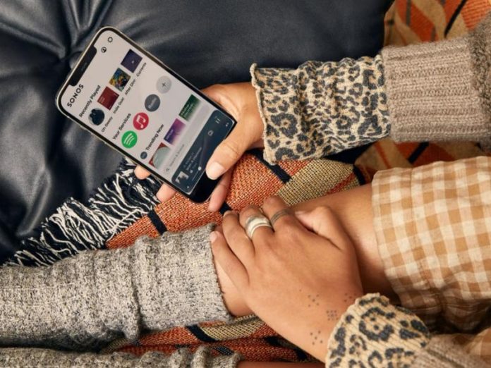 Bovenaanzicht van handen die een goudkleurige smartphone met de openstaande Sonos-app vasthouden. Het scherm toont 'Recently Played' en 'Your Services' met onder andere het Spotify-logo. De handen dragen ringen en hebben kleine tatoeages op de pols. Rondom zijn diverse textielpatronen zichtbaar: zwart leer, een grijze gebreide mouw, een mouw met luipaardprint en een deken met oranje-bruine strepen en ruitpatronen.