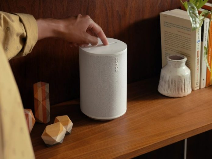 Een hand drukt op de bovenzijde van een witte Sonos-luidspreker op een donkerbruine houten plank, mogelijk om een verbindingsprobleem op te lossen. Rechts staan boeken en een vaas, links decoratieve houten objecten.