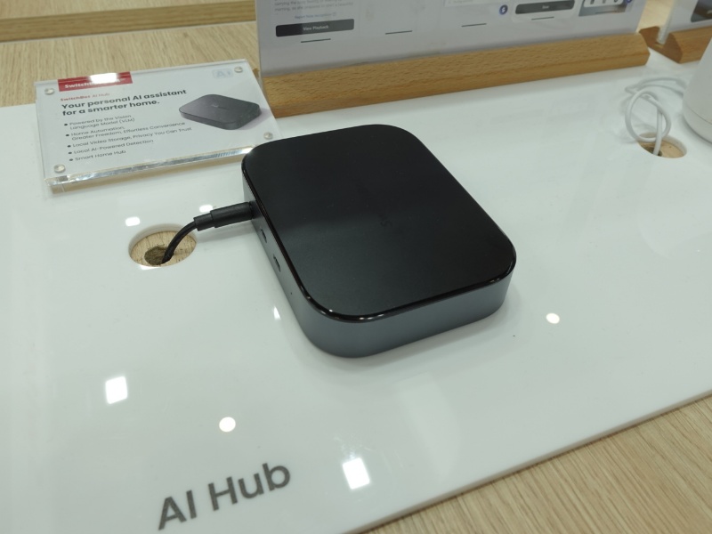 Een zwarte, rechthoekige SwitchBot AI Hub met afgeronde hoeken, via een zwarte kabel aangesloten op een wit displayvlak.