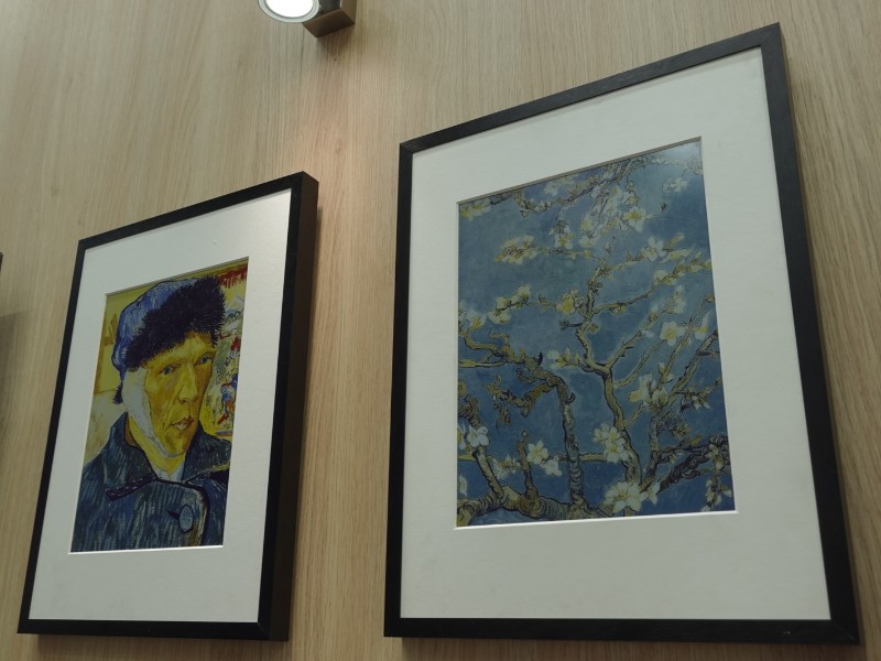 Twee ingelijste Van Gogh Switchbot Ai Frame reproducties hangen aan een lichte houten wand: links een zelfportret met geel gezicht, blauwe muts en verbonden oor, rechts 'Amandelbloesem' met witte bloemen tegen een blauwe lucht. Beide in zwarte lijsten met witte passe-partouts, verlicht door een spot.