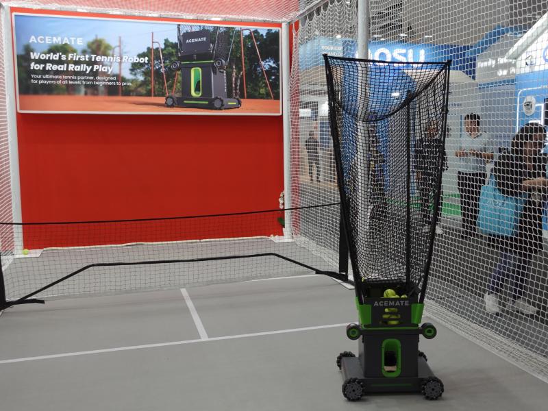 Op een grijze tennisbaan staat een donkergrijze en limoengroene ACEMATE tennisrobot met een zwart opvangnet vol gele tennisballen. Achter de robot, tegen een rode wand, hangt een groot scherm met reclame voor deze 'World's First Tennis Robot'. Rechts zijn door een wit net mensen en een blauwe AOSU stand zichtbaar.