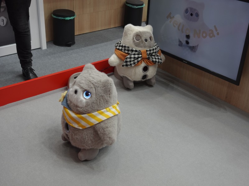 Twee pluizige SwitchBot AI Pets op een grijze vloer: een lichtgrijze met een gloeiend blauw oog en geel-wit gestreepte sjaal staat voorop, terwijl een crèmekleurige met een oranje en zwart-wit geblokte vlinderdas erachter staat. Op een scherm in de achtergrond staat "Hello, Noa!".
