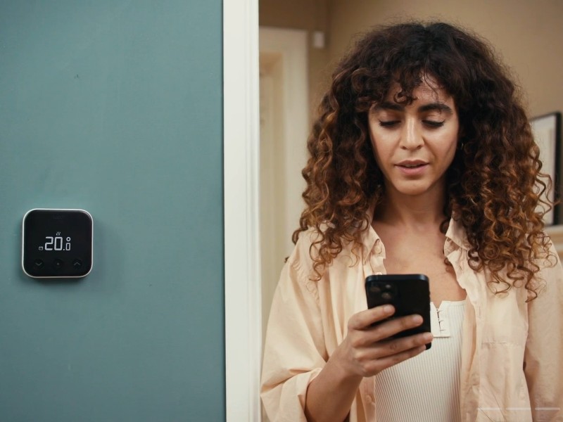 Een vrouw met bruin krullend haar kijkt naar haar smartphone, terwijl links op een petrolblauwe muur een zwarte tado° Thermostaat met wit kader de temperatuur van 20.0° toont.