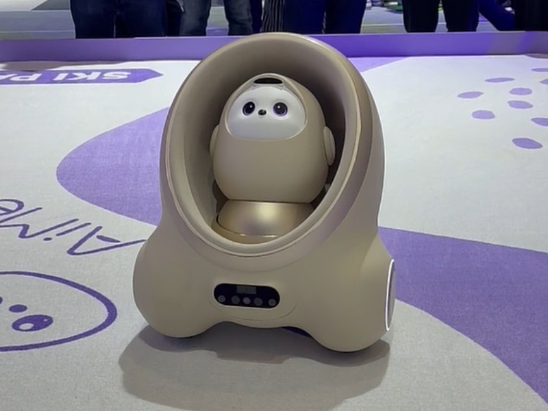 De beige TCL Ai Me robot, met een gouden interieur en daarin een wit robotgezichtje met grote zwarte ogen, staat op een wit tapijt met paarse 'Ai Me' tekst en voetafdrukken. Wazige mensen zijn zichtbaar op de achtergrond.