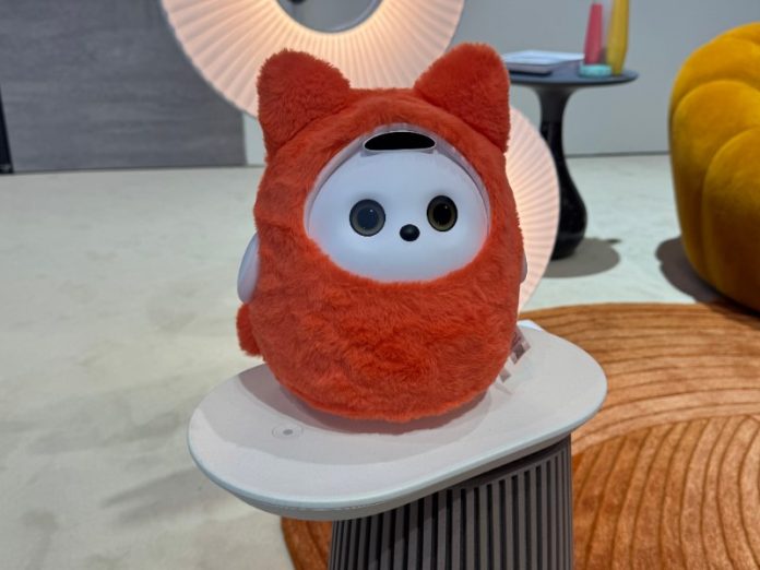 Een pluizige, feloranje TCL Ai Me robot met wit gezicht, grote donkere ogen en een zwarte neus, zit op een lichtgrijs plateau bovenop een donkergrijs geribbeld voetstuk. Op de achtergrond zijn warm verlichte, geplooide ronde lampen, een gele fauteuil en een oranje tapijt zichtbaar.