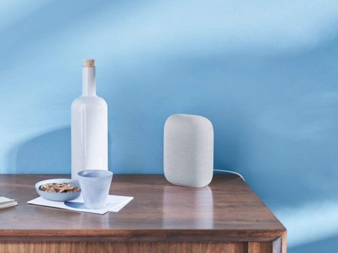 Lichtgrijze Google speaker op houten dressoir voor lichtblauwe muur, naast een witte fles met kurk, een lichtgrijze beker en schaaltje koekjes; passend bij 'Waarom Reageert Mijn Google Speaker Niet? De Oorzaken en Oplossingen'.