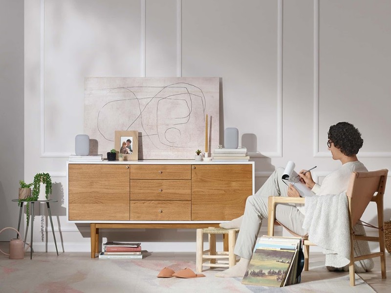 Een persoon met krullend haar en bril zit te schrijven in een lichte, moderne woonkamer. Op een houten dressoir met witte bovenkant staan twee lichtgrijze Google smart speakers, centraal voor de vraag 'Waarom Reageert Mijn Google Speaker Niet? De Oorzaken en Oplossingen'. De minimalistische ruimte heeft witte muren met sierlijsten, een kleurrijk vloerkleed en diverse woonaccessoires zoals planten en vinylplaten.
