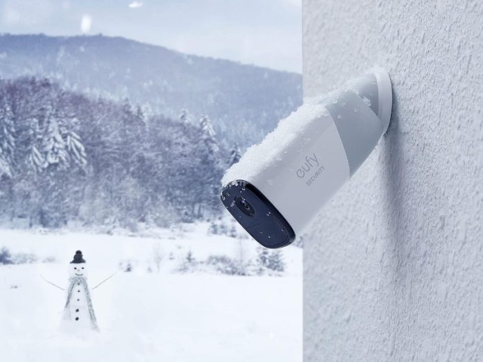 Image van een witte en donkergrijze Eufy beveiligingscamera, bedekt met sneeuw, bevestigd aan een witte, getextureerde muur, met een winters landschap op de achtergrond.