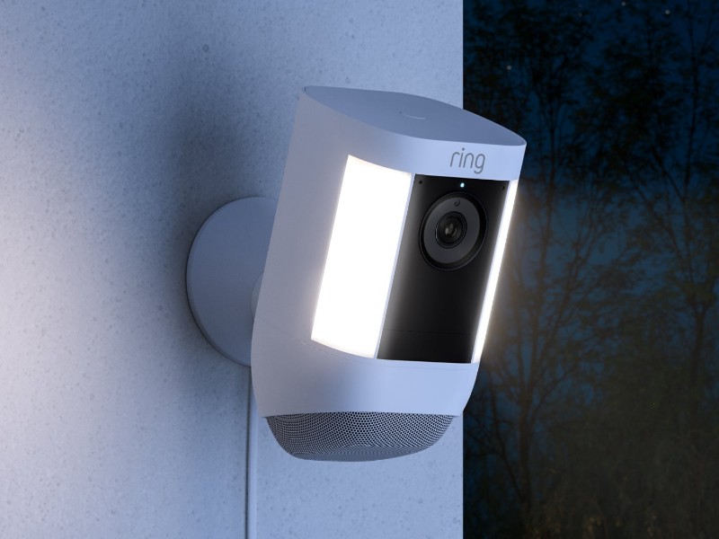 Afbeelding van een witte Ring veiligheidscamera met felle witte LED-spotlights en een zwarte lens, gemonteerd op een lichtgrijze muur tegen een donkere nachtelijke achtergrond.