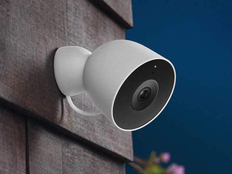 Image van een witte beveiligingscamera met groen lampje, bevestigd aan een donkerbruine houten muur, tegen een donkerblauwe nachthemel met een lichte maan.