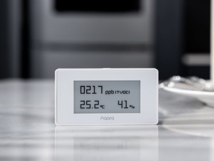 Witte Aqara luchtkwaliteitsmonitor met grijs scherm dat 0217 ppb (TVOC), 25.2°C en 41% vochtigheid toont, staand op een reflecterend lichtgrijs oppervlak voor een wazige donkergrijze achtergrond.