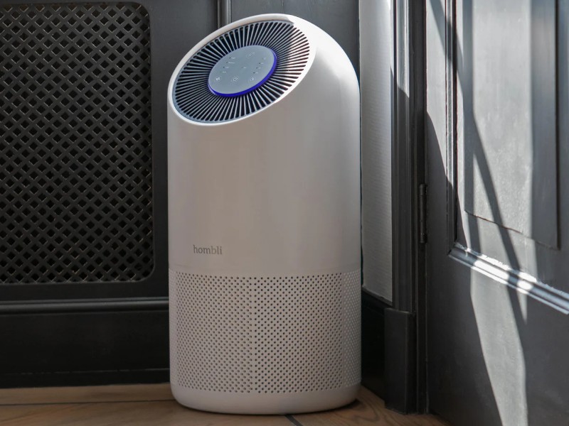 Witte, cilindervormige Hombli luchtreiniger met grijs bedieningspaneel en blauwe verlichting, staand op een lichte houten vloer voor een donkergrijze muur en deur.